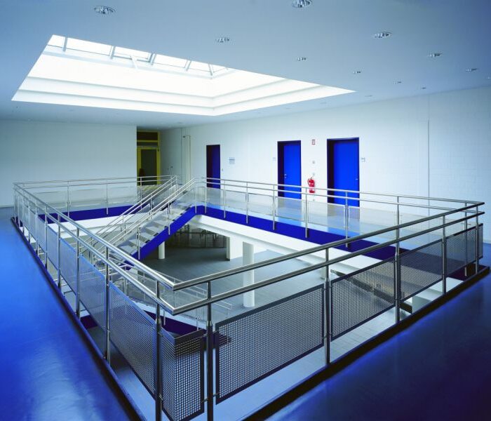 Ein moderner, leerer Innenraum mit blauen Böden, Metallgeländern und einer zentralen Treppe. Mehrere blaue Türen säumen die weißen Wände, und ein großes Oberlicht erhellt den Raum von oben. Ein moderner, leerer Innenraum mit blauen Böden, Metallgeländern und einer zentralen Treppe. Mehrere blaue Türen säumen die weißen Wände, und ein großes Oberlicht erhellt den Raum von oben.