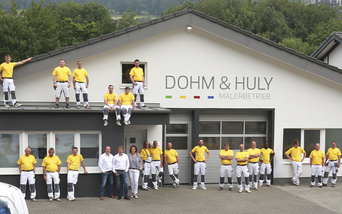 Das Team von Dohm & Huly in gelben Hemden und weißen Arbeitshosen steht und sitzt vor einem Gebäude mit der Aufschrift DOHM & HULY MALERBETRIEB. Einige liegen auf dem Boden, andere sitzen oder stehen auf dem Sims des Gebäudes. Das Team von Dohm & Huly in gelben Hemden und weißen Arbeitshosen steht und sitzt vor einem Gebäude mit der Aufschrift DOHM & HULY MALERBETRIEB. Einige liegen auf dem Boden, andere sitzen oder stehen auf dem Sims des Gebäudes.